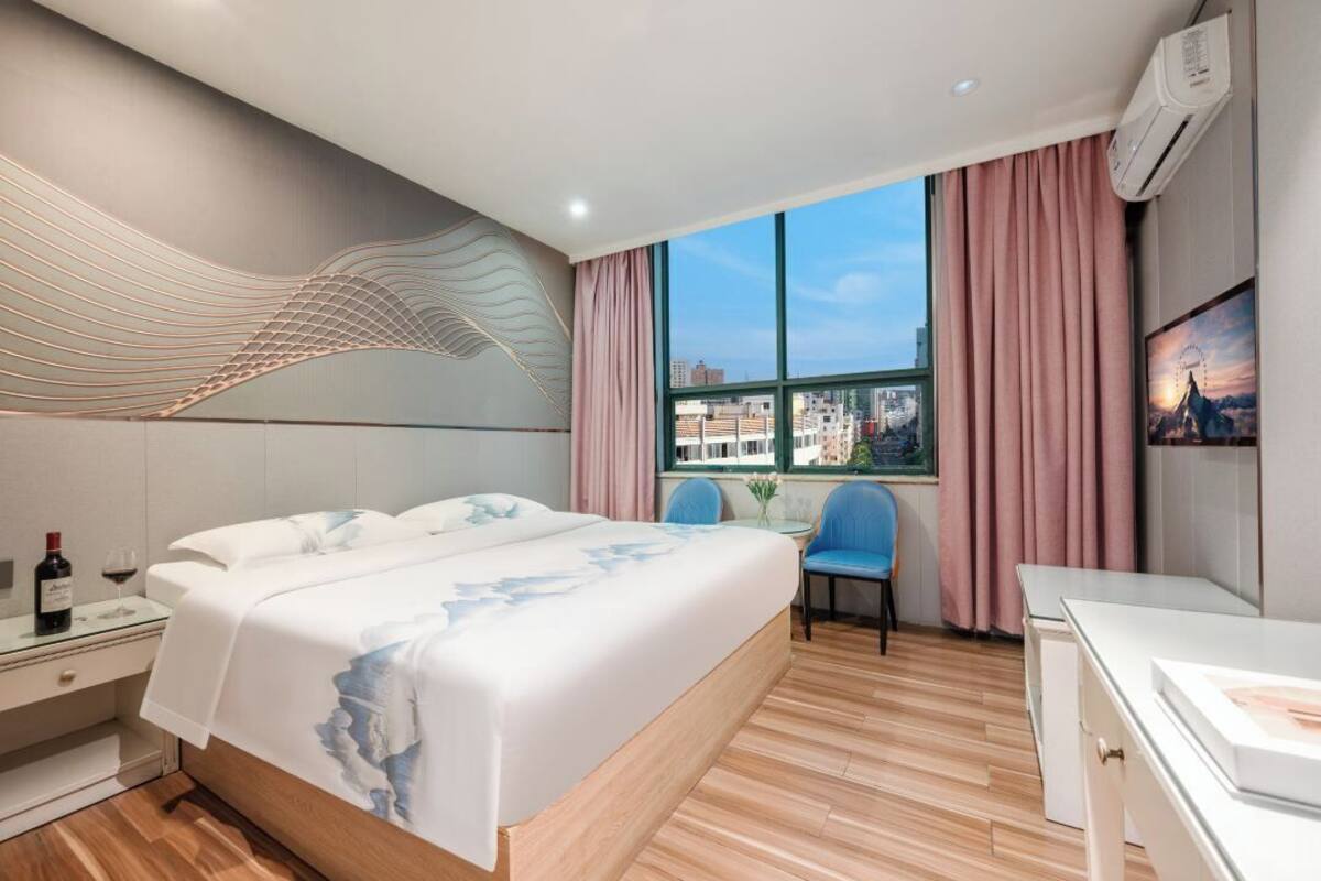 Une chambre d’hôtel moderne avec un grand lit, un bureau et une chaise. Une fenêtre offre une vue sur les bâtiments et le paysage urbain.