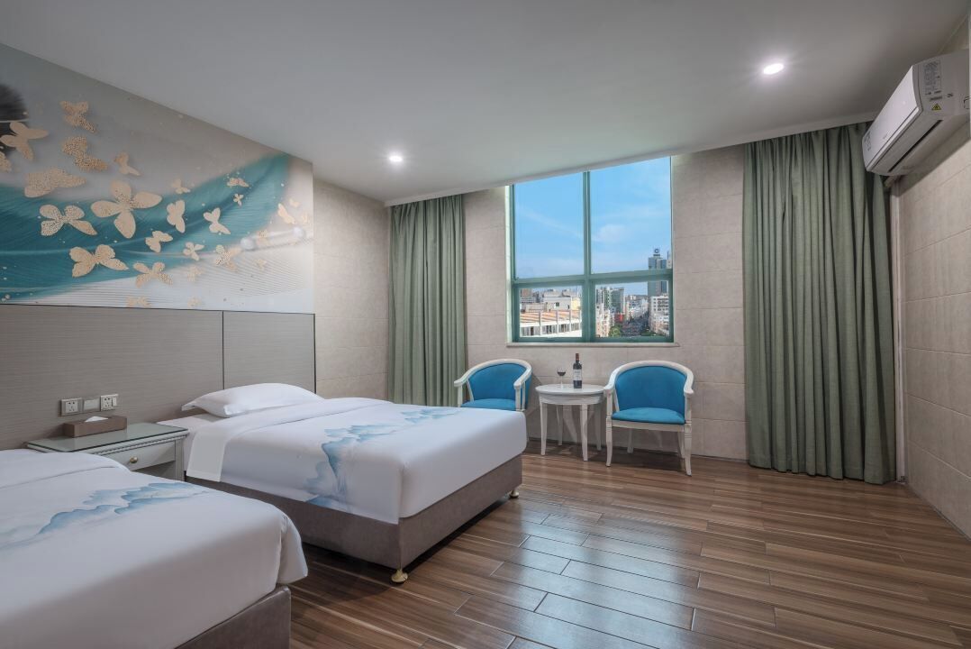 A hotel room with two beds, a small table with chairs, a window with a view of buildings, and an air conditioning unit.