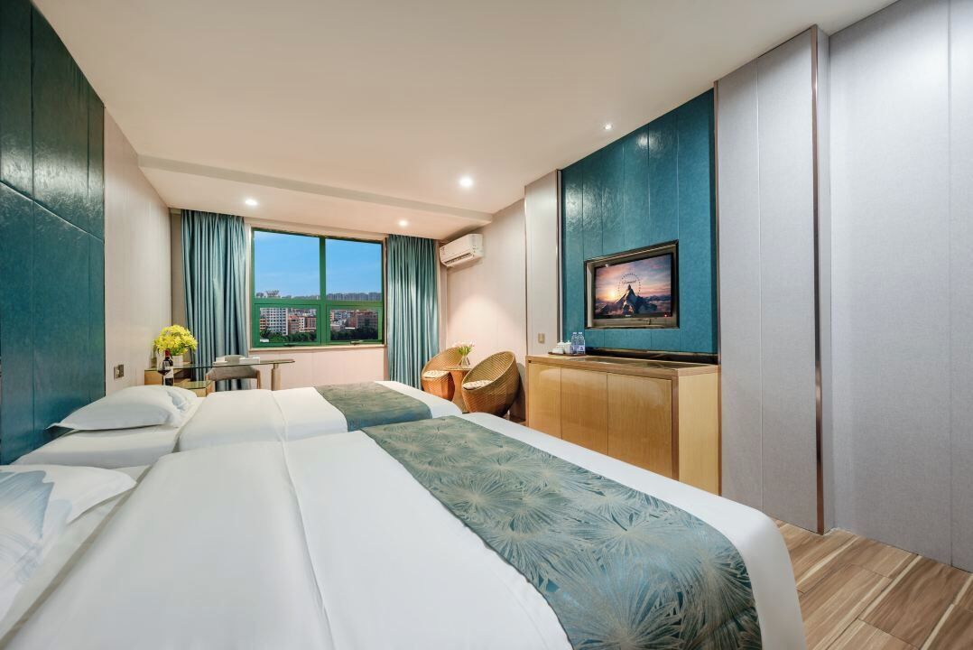 Quarto de hotel com duas camas, uma escrivaninha, uma cadeira, uma televisão e uma janela com vista para a cidade.