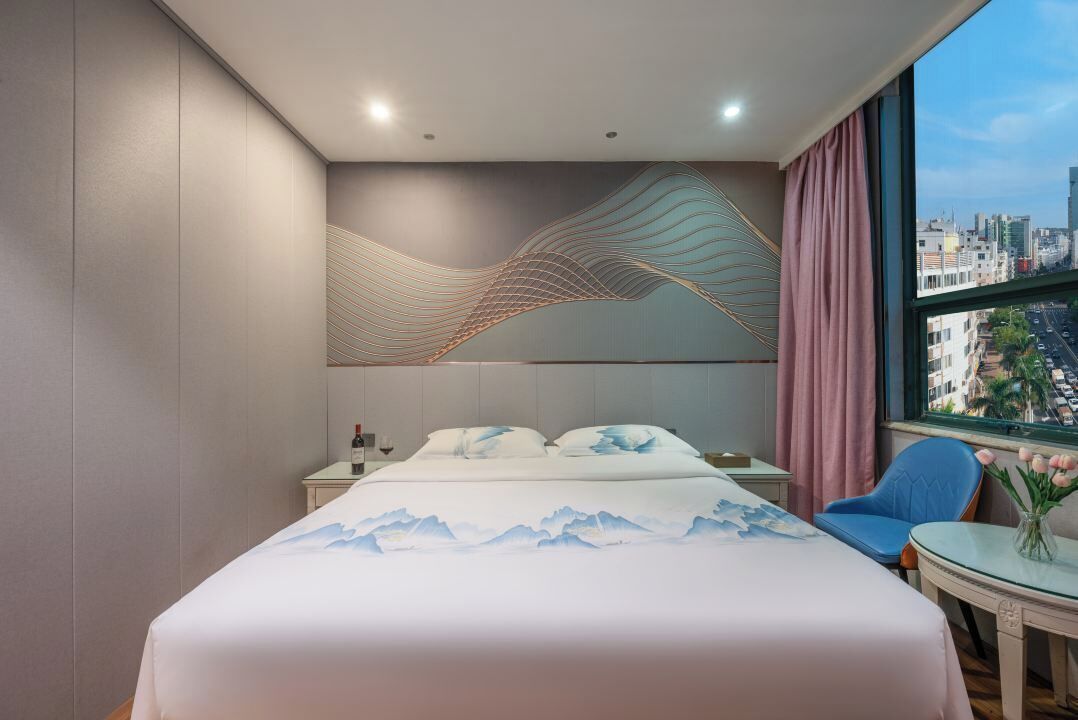A modern hotel room with a bed, a nightstand, a chair, and a view of the city through a large window.
