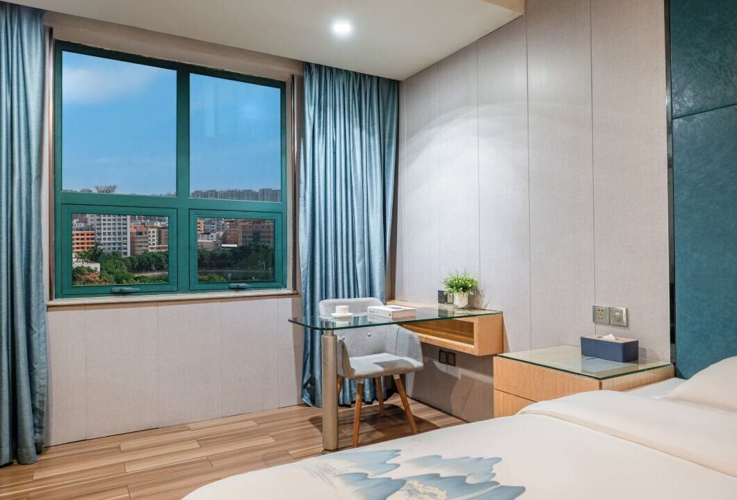 A modern hotel room with a large window, a desk with a chair, a bed, and a small bedside table.
