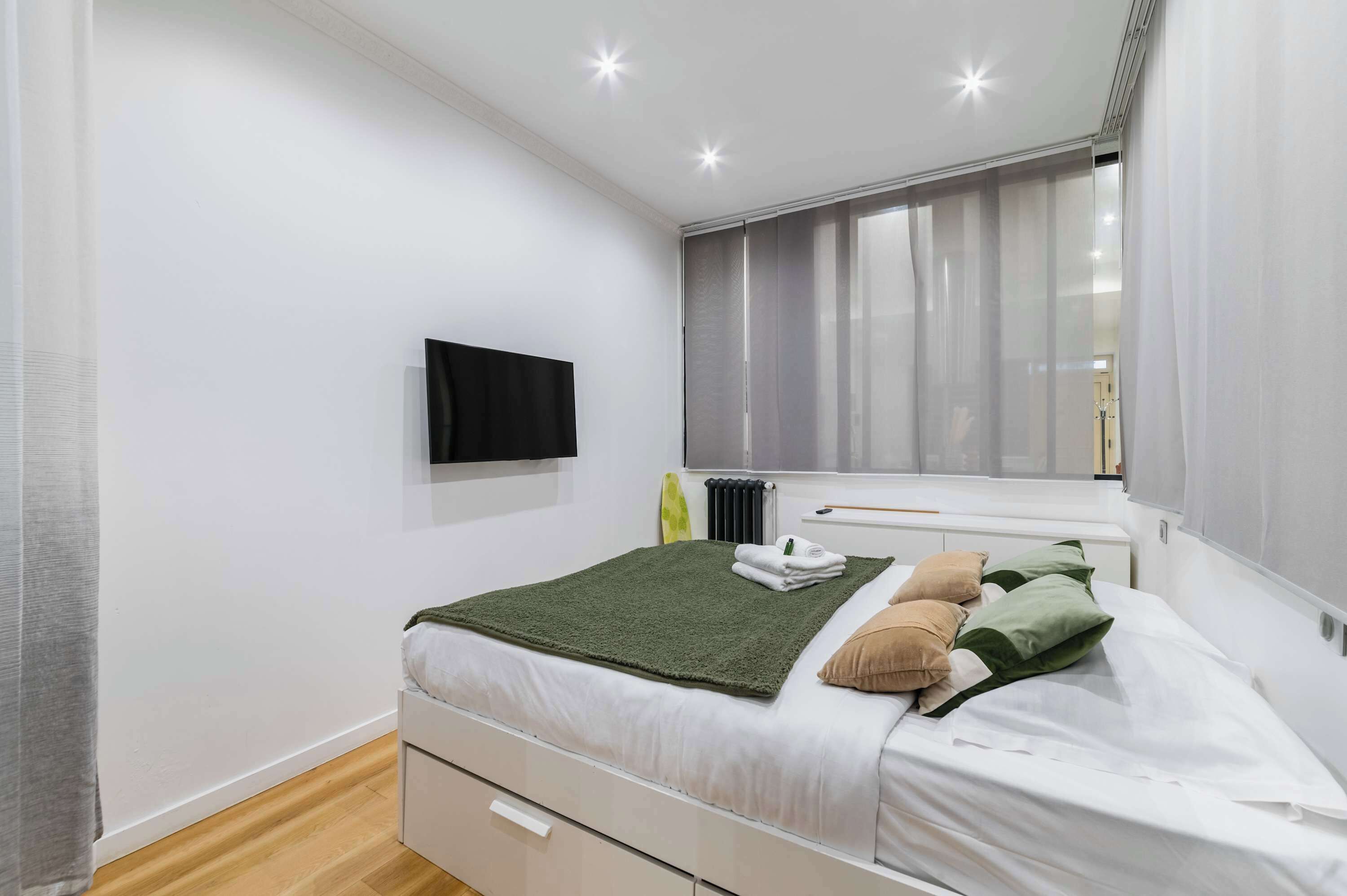 Une chambre à coucher avec un lit, un téléviseur fixé au mur, une fenêtre avec des rideaux et un radiateur.