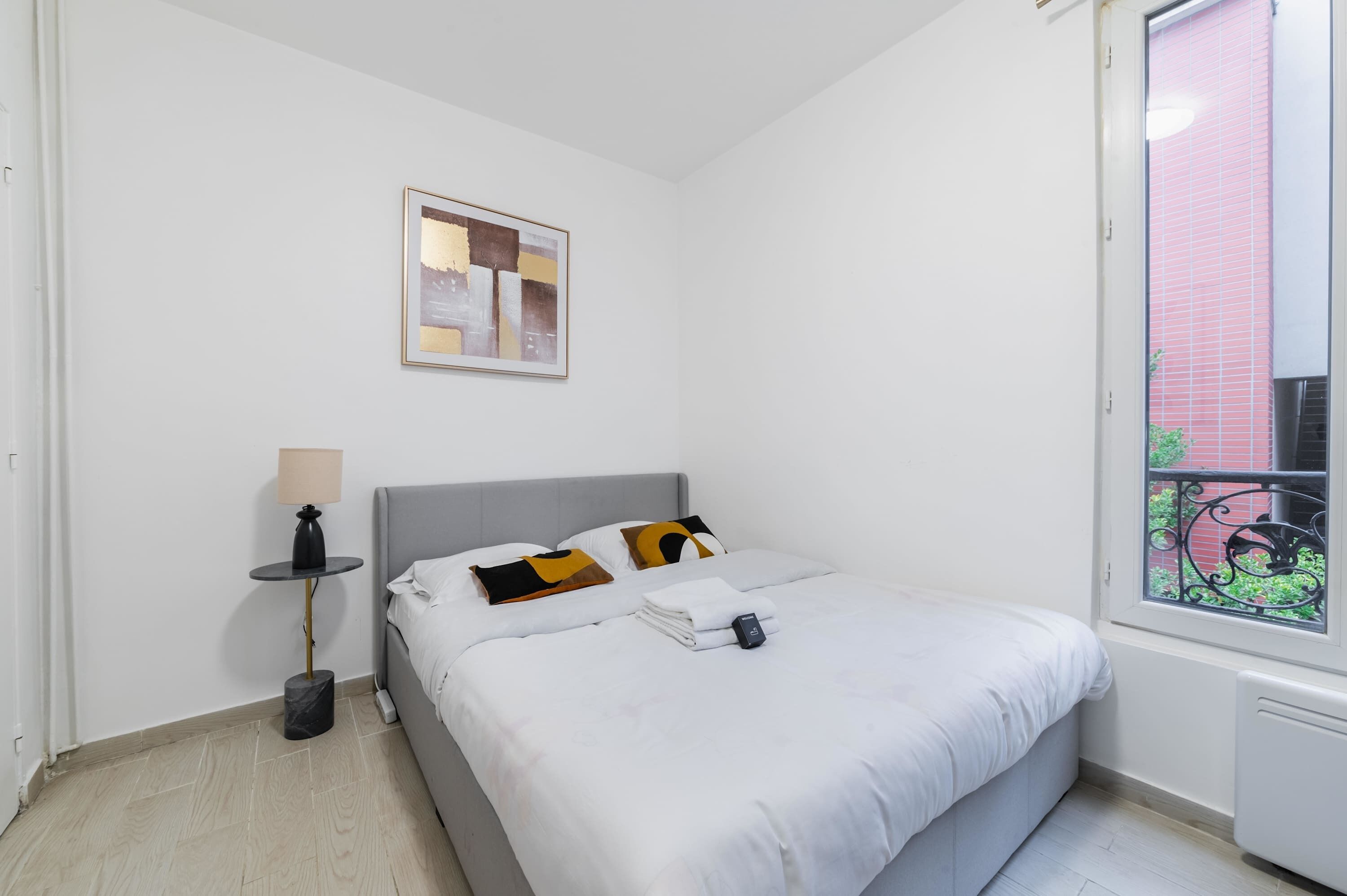 Een netjes opgemaakt bed met wit beddengoed, een nachtkastje met een lamp en een ingelijst beeld aan de muur.
