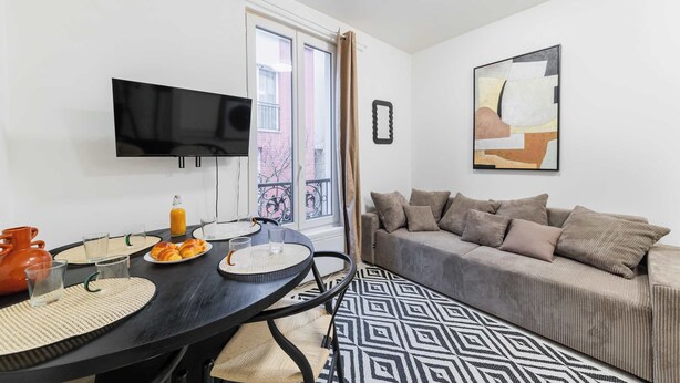 Ein modernes Wohnzimmer mit einer Couch, einem Fernseher, einem Esstisch mit Frühstücksgebäck und einem gemusterten Teppich.