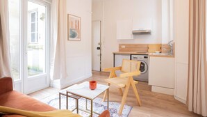 A compact living space with a sofa, a small table, a chair, a kitchen area with a washing machine, and a door leading to an outdoor area. - Logement cosy metro Pierre Lachaise - 3P (Paris)