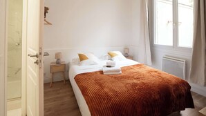 A bedroom with a bed, a nightstand, a lamp, a window, and a door. - Logement cosy metro Pierre Lachaise - 3P (Paris)