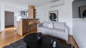 A modern living room with a sofa, a coffee table, a remote control, and a kitchen area with a counter and chairs. - Appartement lumineux avec terrasse Courbevoie - 4P (Courbevoie)