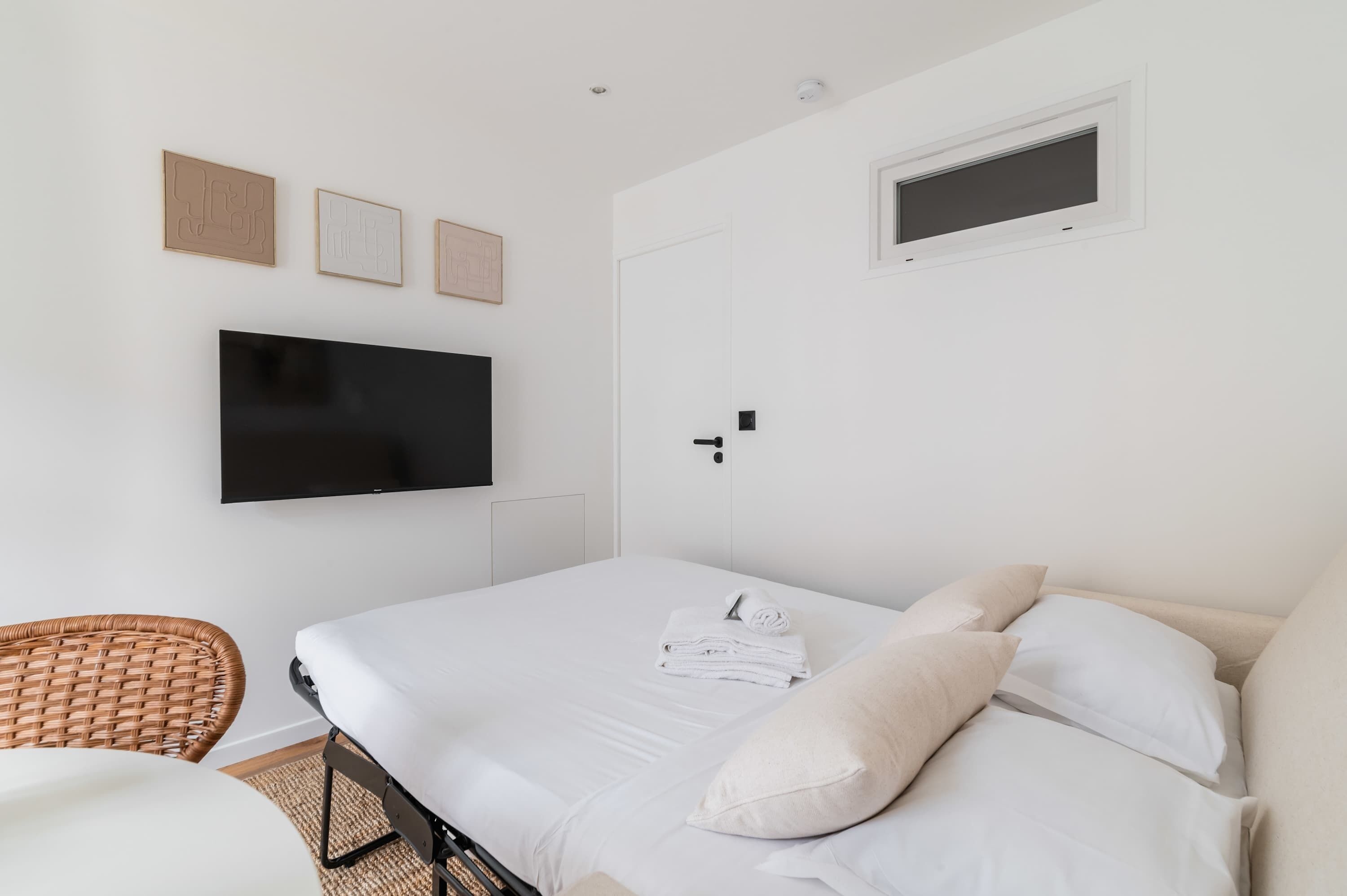 A hotel room with a bed, a flat-screen TV, a wall-mounted light switch, and a small window.