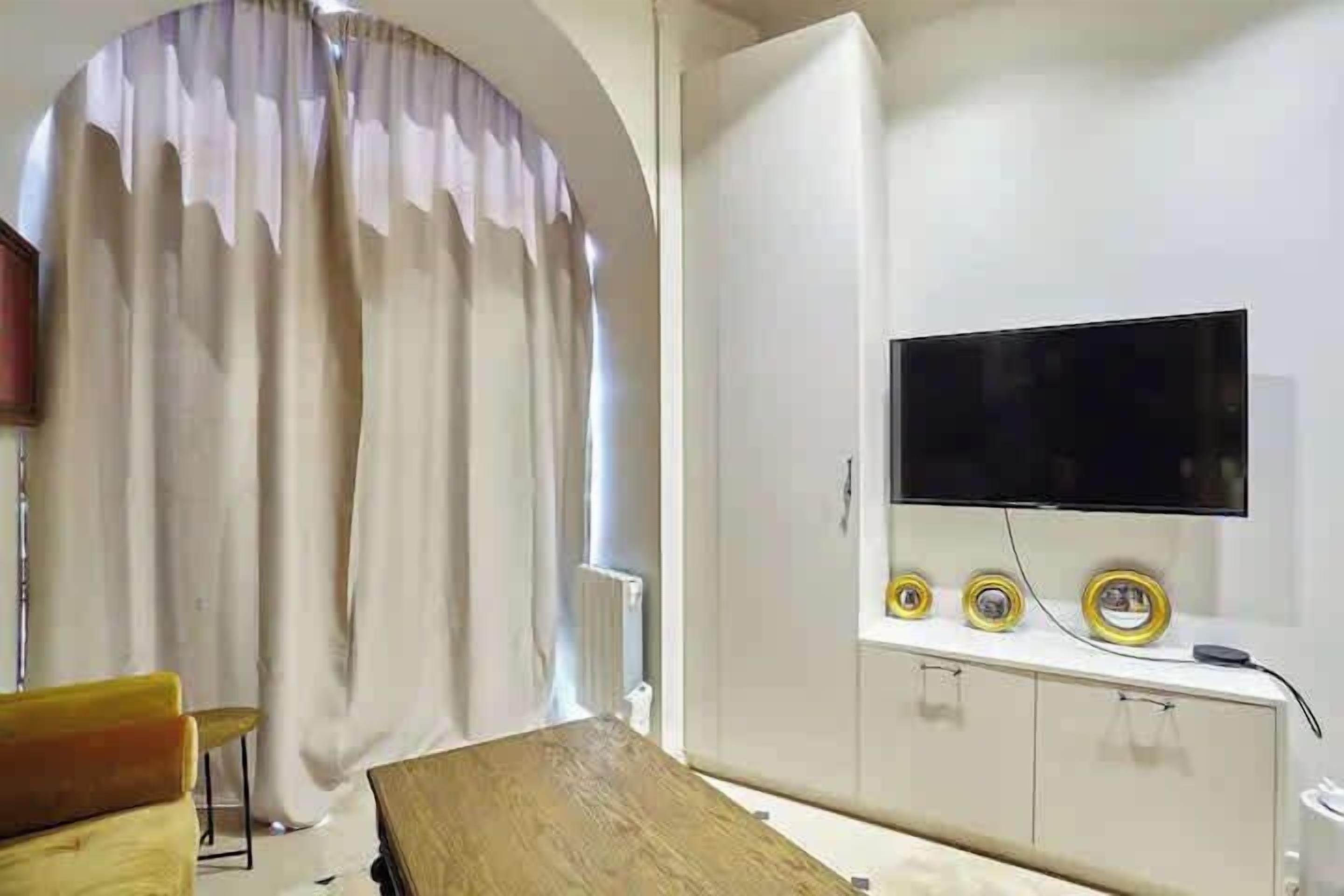 A room with a TV, a wooden table, a chair, a window with curtains, and a white wardrobe.