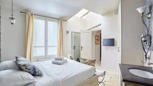 A hotel room with a bed, a bedside table, a mirror, a window, and a bathroom with a sink and mirror.