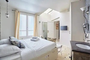 A hotel room with a bed, a bedside table, a mirror, a window, and a bathroom with a sink and mirror.