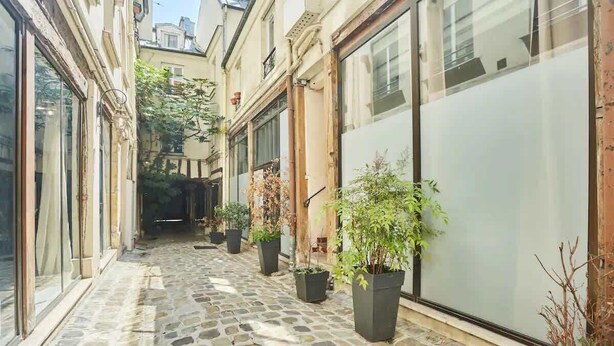 A narrow stone-paved courtyard with potted plants, large windows, and a view of the street.