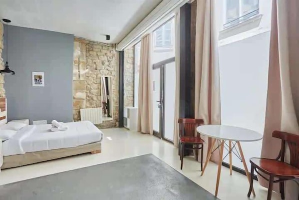 A hotel room with a bed, a small table, chairs, a stone accent wall, and a view through a window.