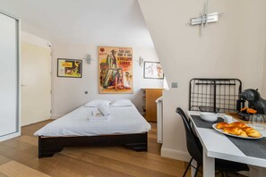 A bedroom with a bed, a dining table with croissants, a kettle, and a poster on the wall. - Studio refait a neuf paris 17 climatisation - 2P (Paris)