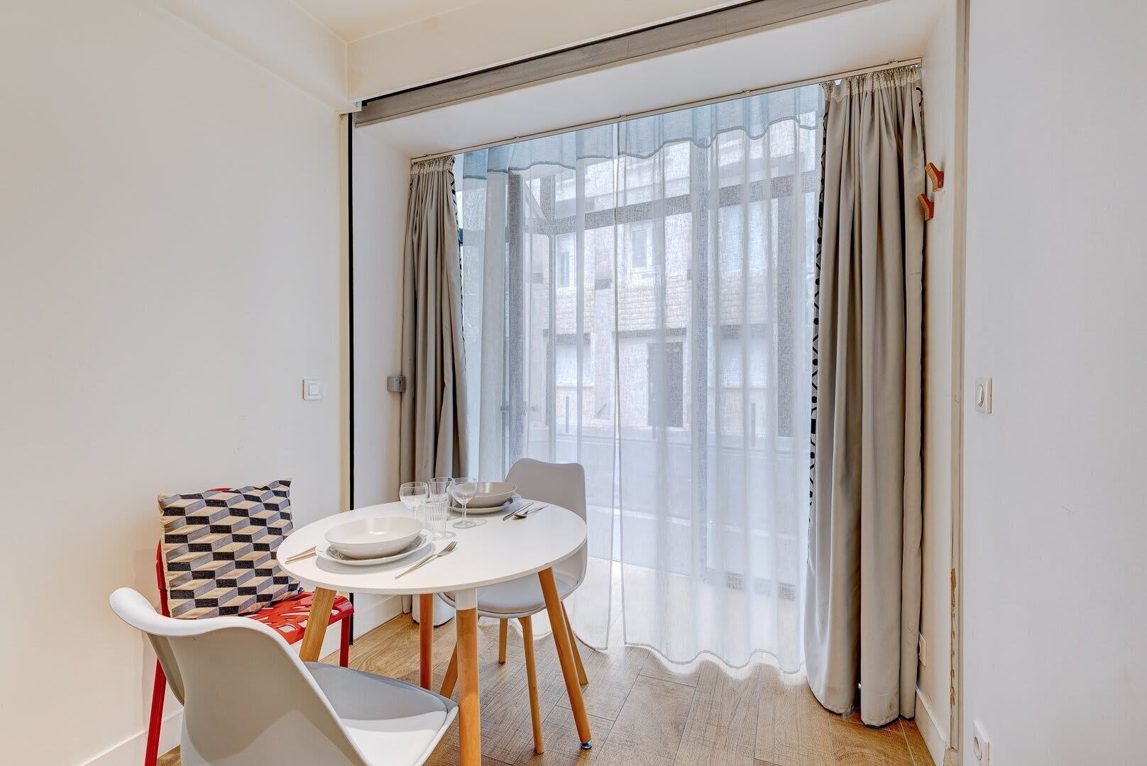 A dining area with a round table, chairs, and a view through sheer curtains.