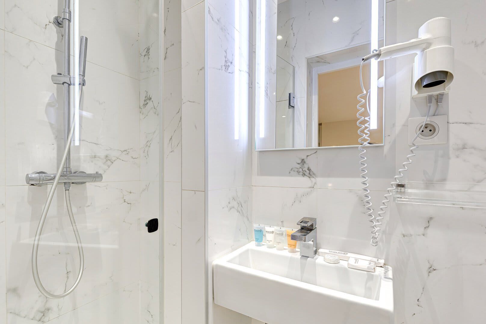 A modern bathroom with a marble shower, a sink with toiletries, and a hairdryer.