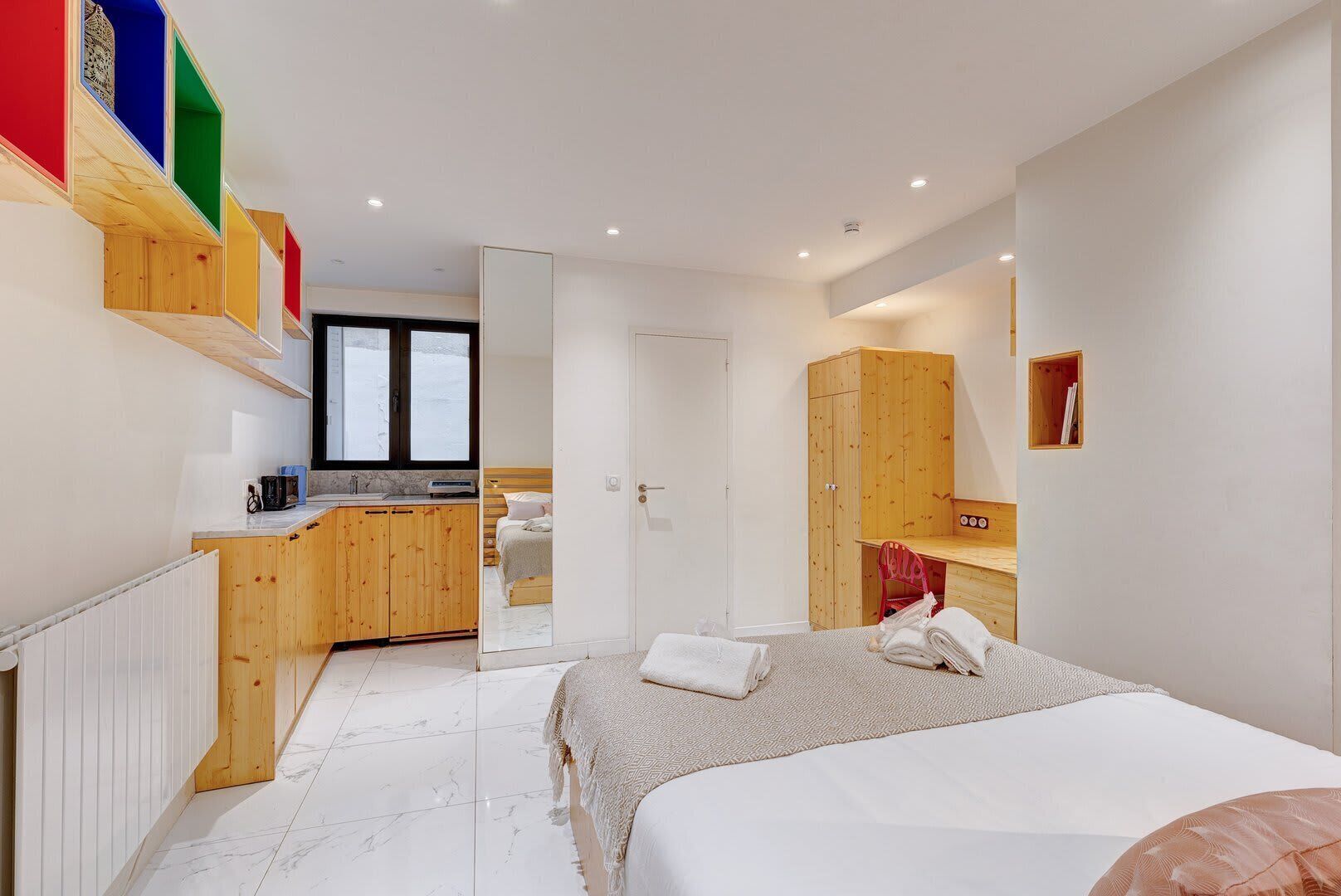 A bedroom with a bed, a wooden wardrobe, a desk, and a window.
