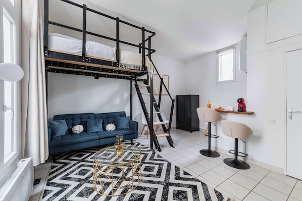 Una habitación con literas, un sofá azul, dos taburetes altos y una pequeña cocina con cafetera.