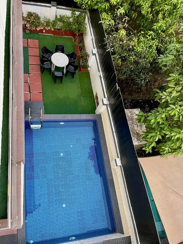 A rectangular swimming pool with a tiled floor, surrounded by a low brick wall and a green lawn area with a small table and chairs. - Greystone 4BHK Pvt Pool by Stellar Stays (Mawal)
