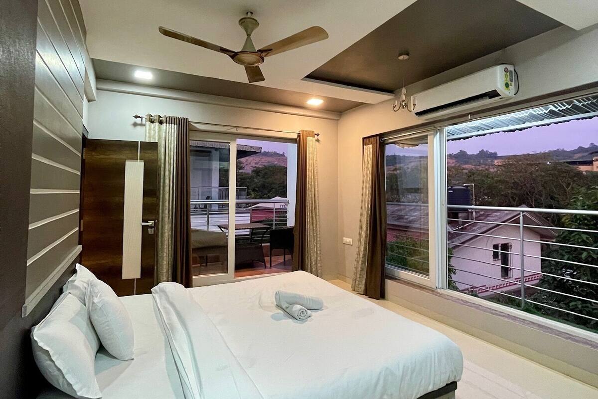 A modern bedroom with a bed, ceiling fan, air conditioning unit, and large windows offering a view of the outdoors.