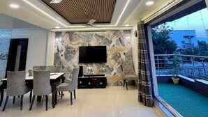 A modern living room with a flat-screen TV, a dining area with chairs, a ceiling fan, and a large window with a view of the outdoors. - Greystone 4BHK Pvt Pool by Stellar Stays (Mawal)