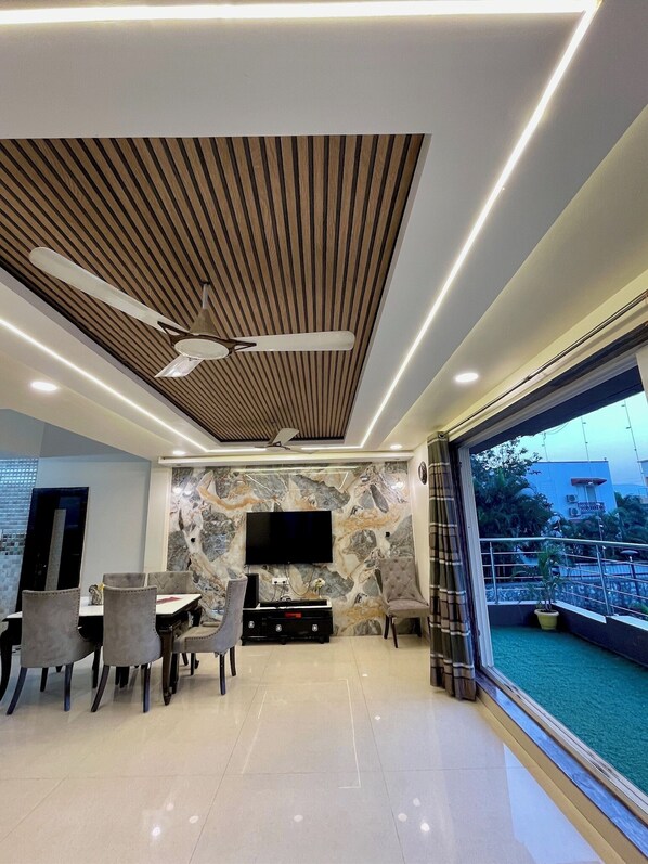 A modern living room with a flat-screen TV, a dining area with chairs, a ceiling fan, and a large window with a view of the outdoors. - Greystone 4BHK Pvt Pool by Stellar Stays (Mawal)