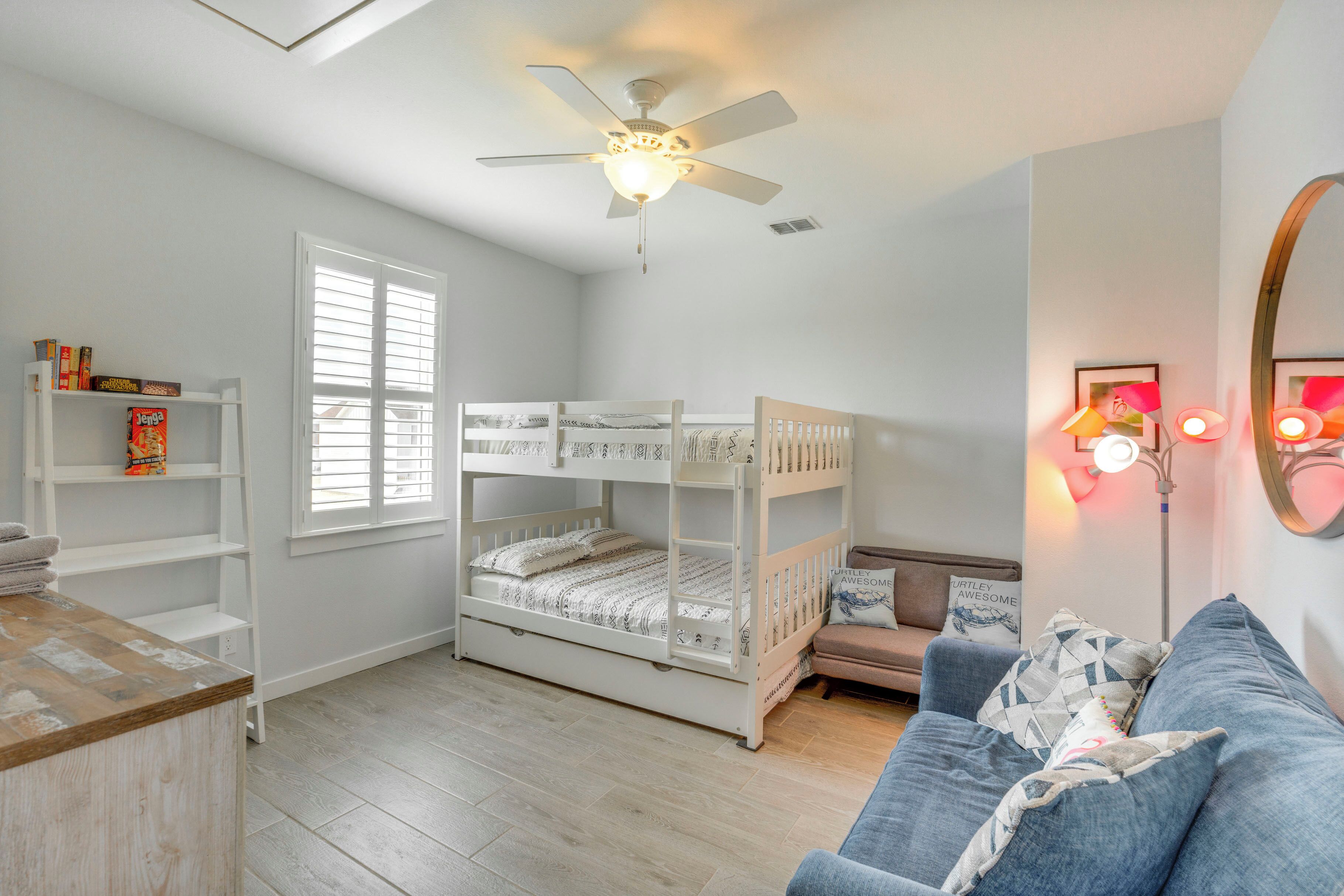 Une chambre avec un lit superposé, un canapé, un ventilateur de plafond, une fenêtre avec des stores et une étagère remplie de livres.