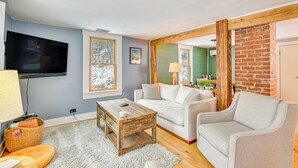 Ein Wohnzimmer mit einem Flachbildfernseher, einem Holztisch, einem weißen Sofa und einem grauen Sessel. Im Hintergrund sind eine Ziegelwand und eine grüne Wandverkleidung zu sehen.