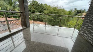 Uma varanda com guarda-corpo de vidro, piso de azulejos e vista para palmeiras e um céu nublado.