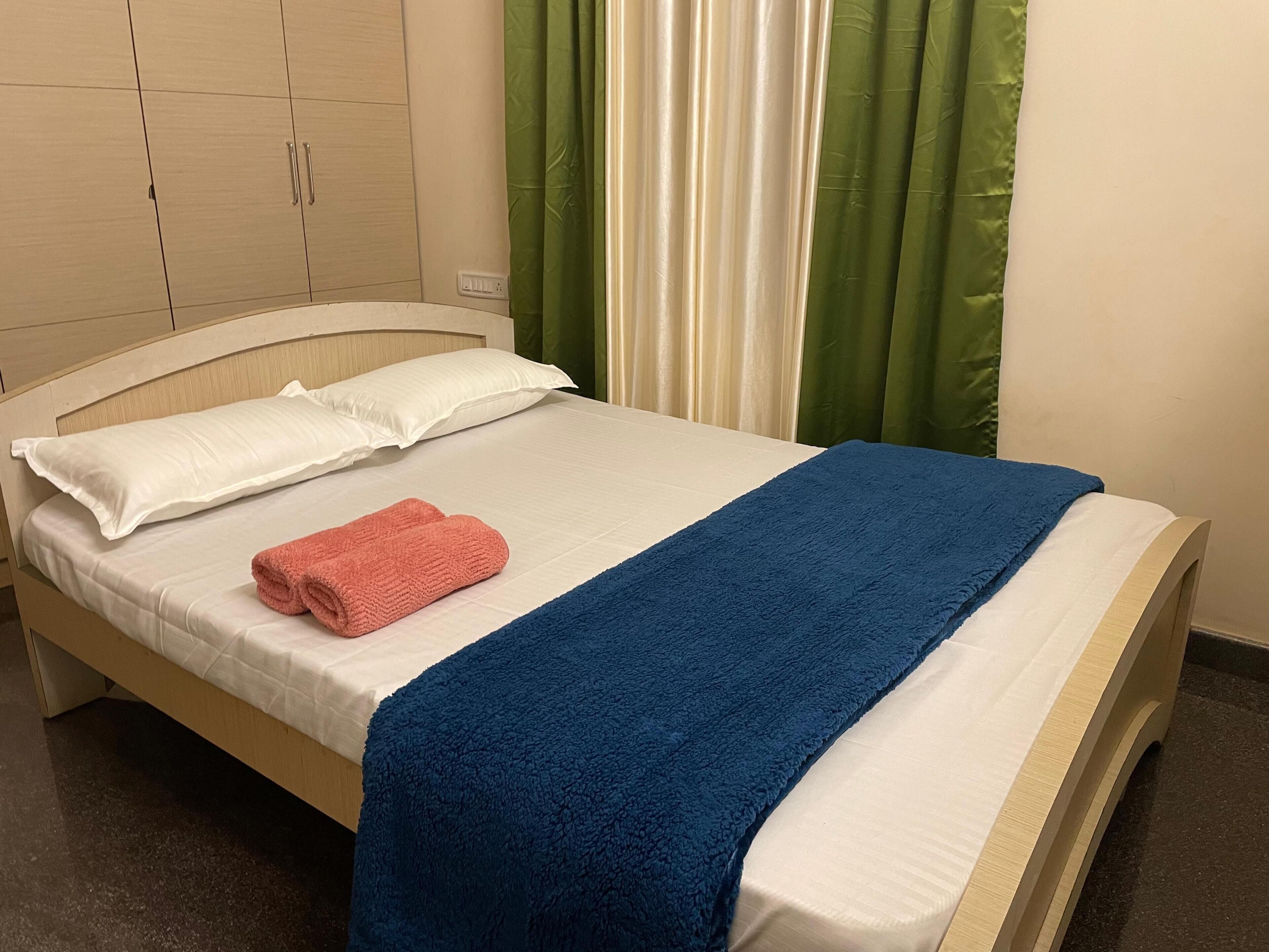 Una cama bien hecha con ropa de cama blanca y azul, una toalla rosa y cortinas verdes al fondo.