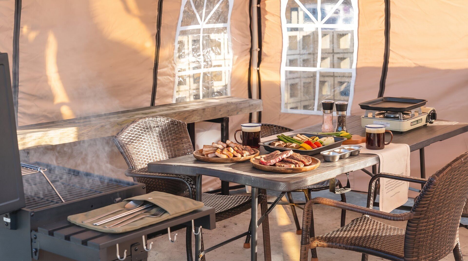 Un área de comedor al aire libre con carpas, una parrilla, sillas de mimbre y una mesa servida con comida y bebidas.