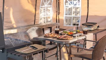Un área de comedor al aire libre con carpas, una parrilla, sillas de mimbre y una mesa servida con comida y bebidas.