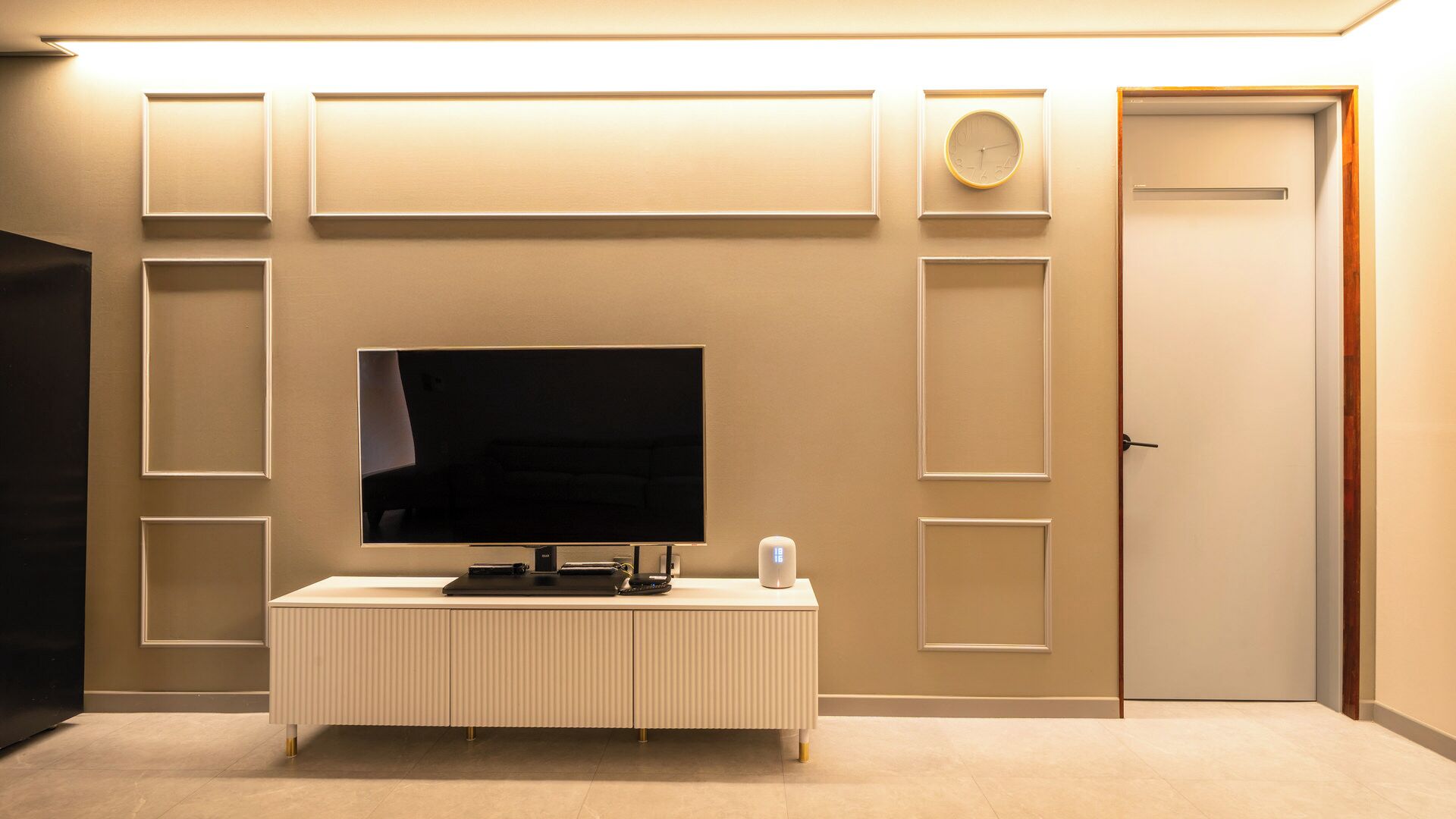 Une pièce avec un téléviseur à écran plat posé sur une console blanche, une porte et des horloges fixées au mur.