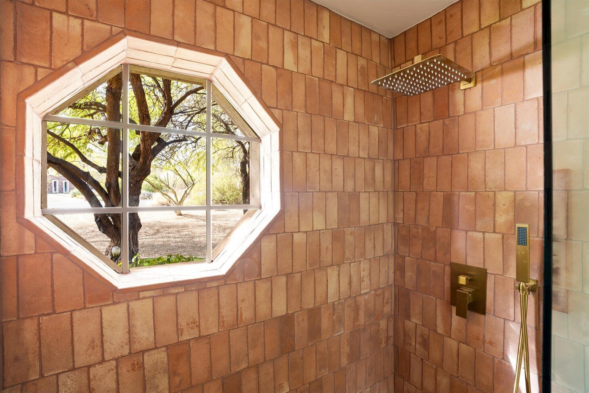 A bathroom with a large octagonal window, a shower with a gold showerhead, and a tiled wall.