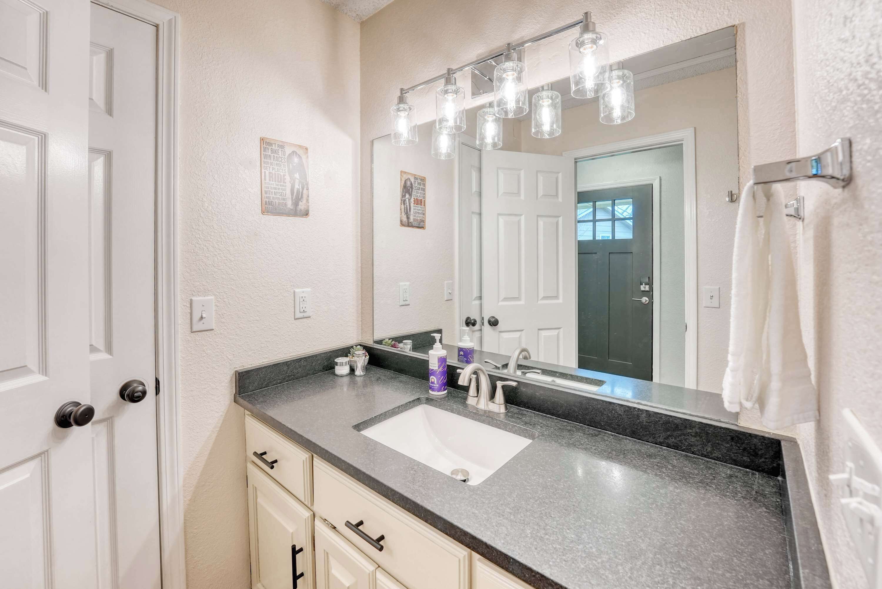 Une salle de bain avec un lavabo blanc, un grand miroir et un plan de toilette sur lequel se trouvent un distributeur de savon et une bouteille.