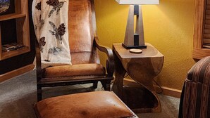 Ein gemütliches Zimmer mit einem Holzstuhl und einem Hocker, einem kleinen Tisch mit einer Lampe und einem Regal mit Büchern und einem gerahmten Bild.