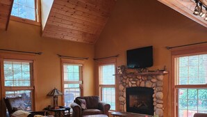 Ein geräumiges Wohnzimmer mit Holzböden, einem Kamin aus Stein, Ledersofas und großen Fenstern.