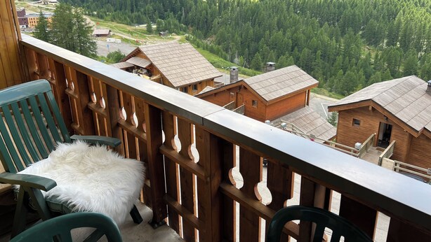A wooden balcony with a view of a forested valley, a rustic building, and a chair with a fluffy cushion.