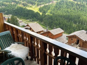 A wooden balcony with a view of a forested valley, a rustic building, and a chair with a fluffy cushion. - Magnificent apartment right in the center, walking distance to shops and ski slopes! Terrace (Isola)