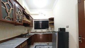 A compact kitchen with a marble countertop, wooden cabinets, a refrigerator, and a black window blind. - Room-serene Retreat in Rawalpindi - Your (Rawalpindi)