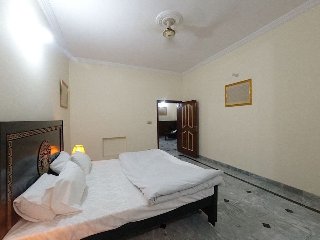 A hotel room with a bed, a nightstand, a lamp, and a door leading to another room.