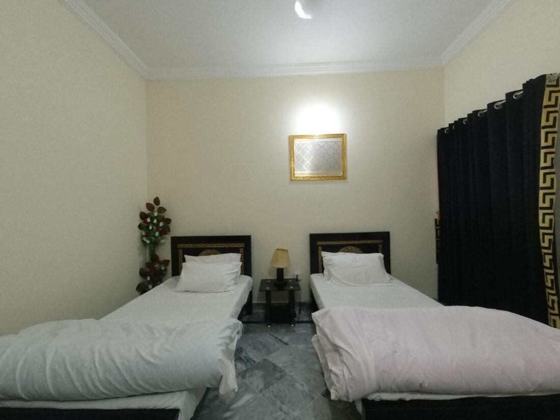 Habitación de hotel con dos camas individuales, una mesita de noche con lámpara, un cuadro en la pared y una cortina con estampado geométrico.