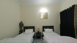 Une chambre d’hôtel avec deux lits simples, une table de chevet avec une lampe, un tableau encadré au mur et un rideau à motifs géométriques.