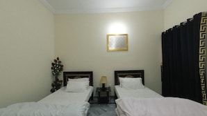 Une chambre d’hôtel avec deux lits simples, une petite table avec une lampe, un tableau encadré au mur et un rideau à motifs géométriques.
