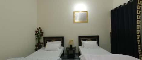 A hotel room with two single beds, a nightstand with a lamp, a framed picture on the wall, and a curtain with a geometric pattern.