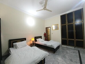 A hotel room with two beds, a ceiling fan, a nightstand with a lamp, a large window, and a wardrobe. - Room-cozy Guest Room in Rawalpindi - Per (Rawalpindi)