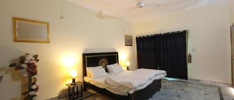 A hotel room with a large bed, a ceiling fan, a decorative vase with flowers, and a dark curtain on the window.