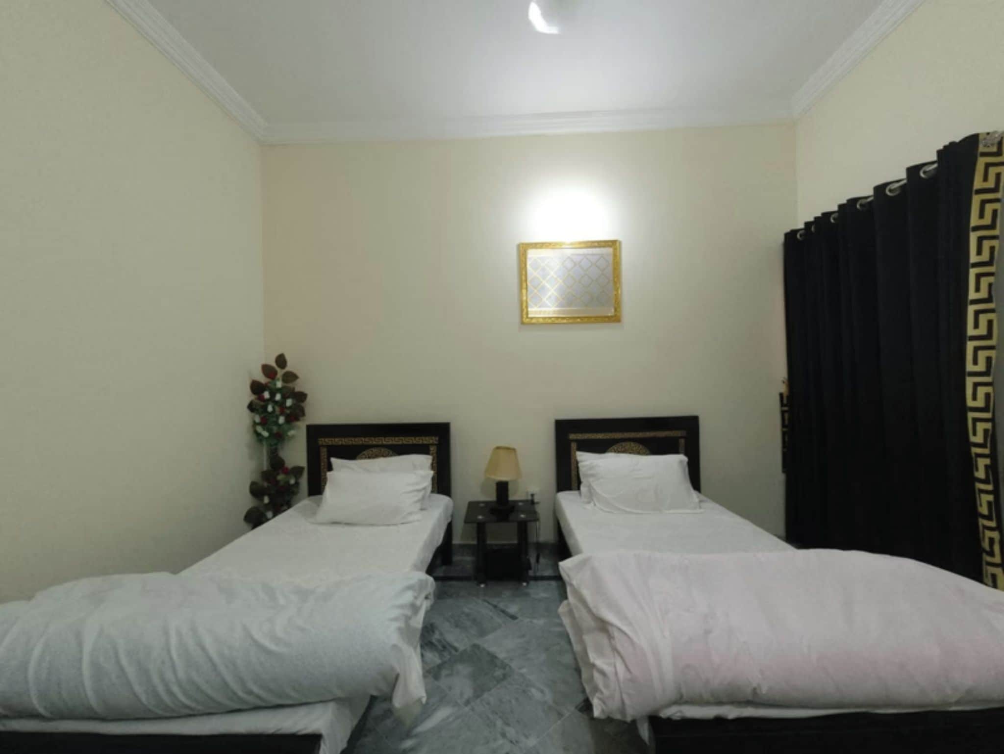 A hotel room with two single beds, a nightstand with a lamp, a framed picture on the wall, and a curtain with a geometric pattern.