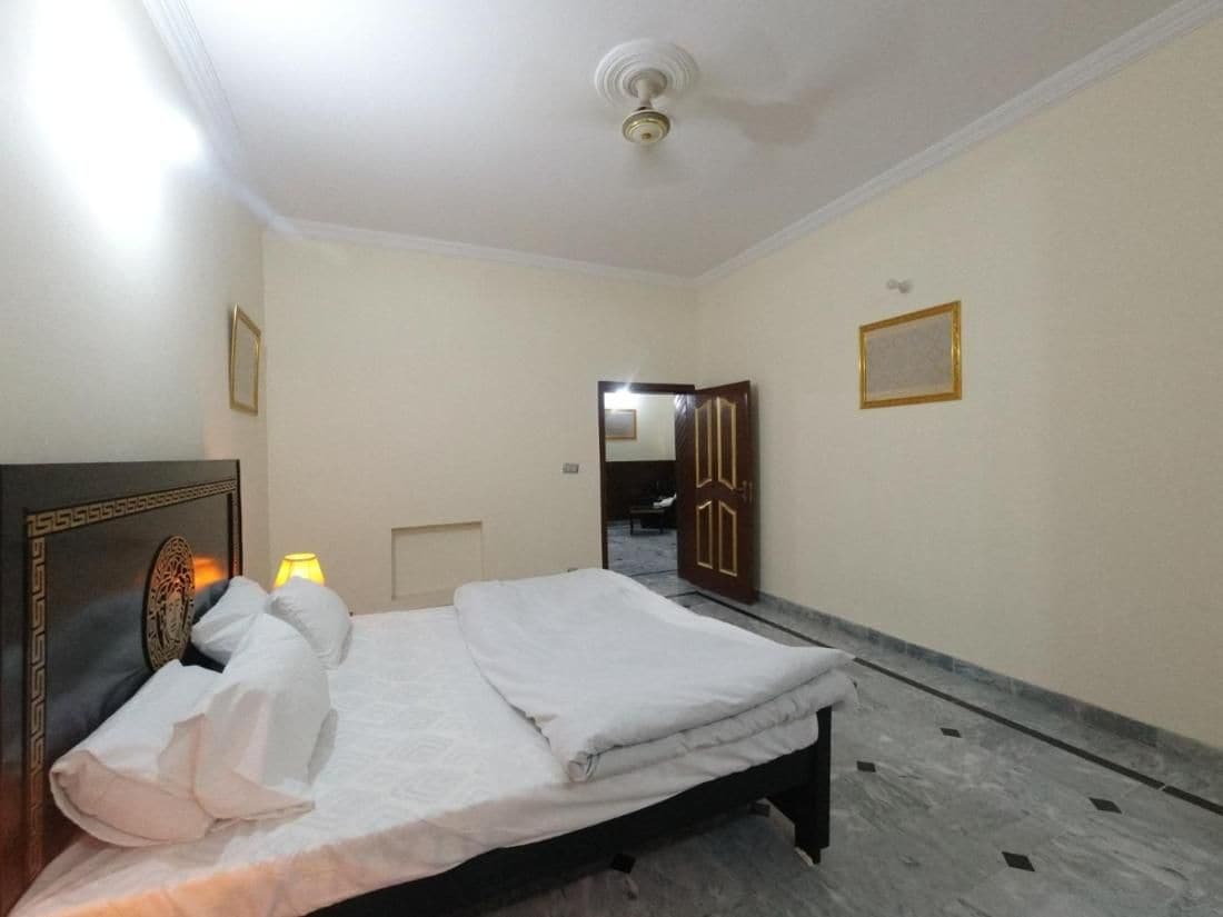 A hotel room with a bed, a nightstand, a lamp, and a door leading to another room.