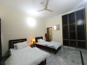 A hotel room with two beds, a ceiling fan, a nightstand with a lamp, a large window with a view, and a wardrobe. - Room-cozy Guest Room in Rawalpindi - Per (Rawalpindi)